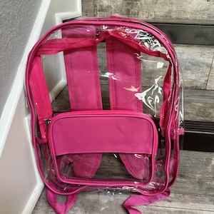 May include: A clear pink backpack with a pink fabric front pocket. The backpack has adjustable straps and a zipper closure.