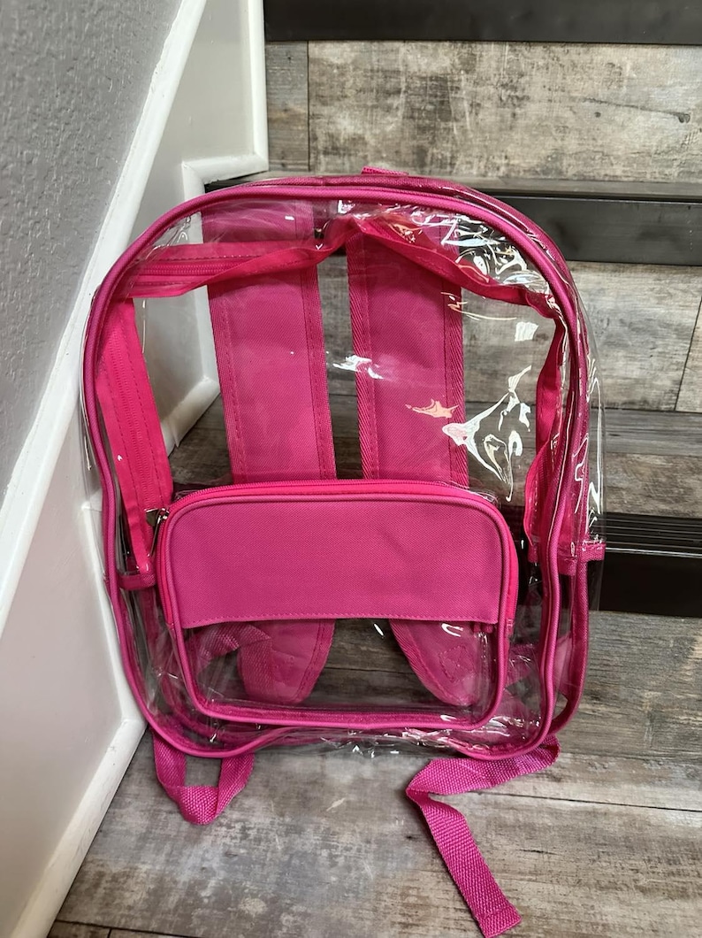 May include: A clear pink backpack with a pink fabric front pocket. The backpack has adjustable straps and a zipper closure.