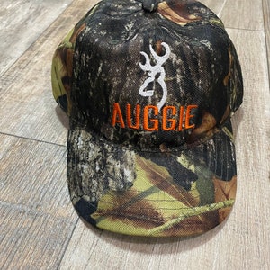 May include: Camouflage baseball cap with the word "AUGGIE" in orange letters. The cap features a white emblem and a pattern of brown, green, and yellow. The cap is made of a durable fabric.