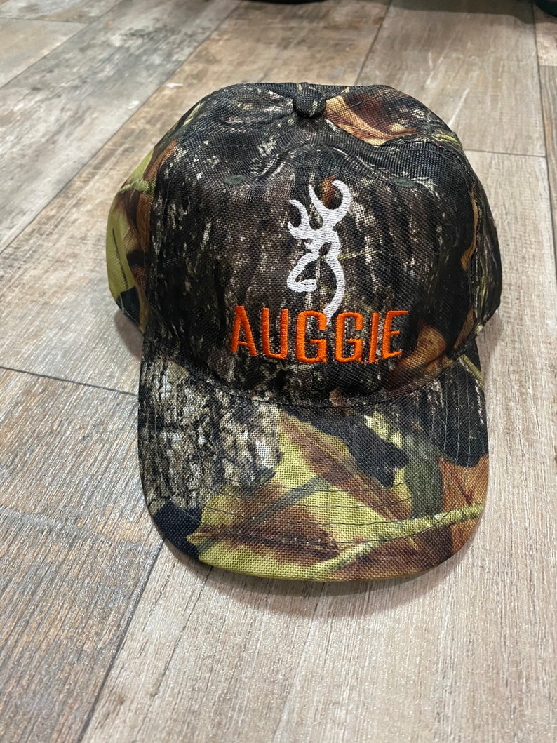 May include: Camouflage baseball cap with the word "AUGGIE" in orange letters. The cap features a white emblem and a pattern of brown, green, and yellow. The cap is made of a durable fabric.