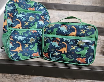 May include: A navy blue backpack and a matching rectangular lunch bag, both featuring a colorful dinosaur pattern with green, orange, and light blue dinosaurs. The lunch bag has a green handle and a small white label reading Stephen Joseph.