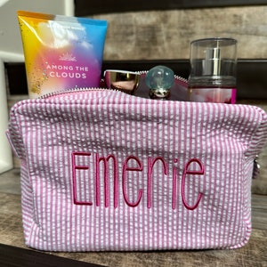 May include: Pink and white striped zippered toiletry bag with the name "Emerie" embroidered in pink on the front. The bag is filled with various toiletries, including a bottle of body lotion, a bottle of perfume, and a pair of sunglasses.