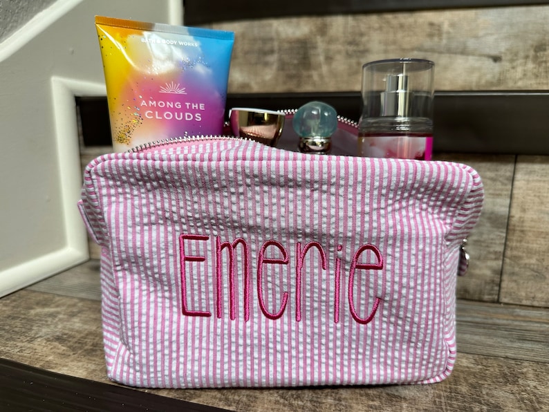 May include: Pink and white striped zippered toiletry bag with the name "Emerie" embroidered in pink on the front. The bag is filled with various toiletries, including a bottle of body lotion, a bottle of perfume, and a pair of sunglasses.