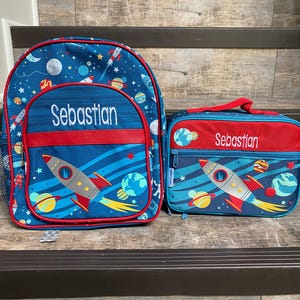 May include: A blue and red space-themed backpack and lunch bag set with a rocket ship design. The backpack and lunch bag are personalized with the name "Sebastian".