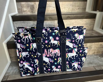 May include: A navy blue tote bag with a unicorn print and the name "Alora" embroidered on the front. The bag has multiple pockets and a zipper closure.