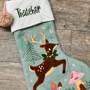 May include: A light green Christmas stocking with a white cuff embroidered with the name Thatcher in dark green. The stocking features an applique of a brown deer with gold antlers, green pine trees, gold stars, and pink and brown mushrooms.