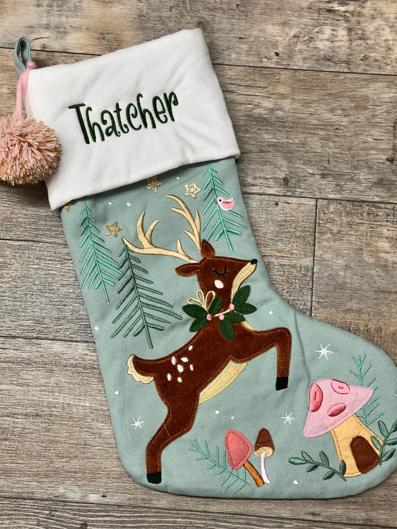 May include: A light green Christmas stocking with a white cuff embroidered with the name Thatcher in dark green. The stocking features an applique of a brown deer with gold antlers, green pine trees, gold stars, and pink and brown mushrooms.
