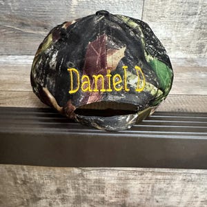 May include: A camouflage baseball cap with a yellow embroidered name "Daniel D" on the front.