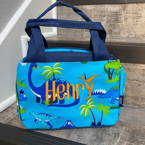 May include: Blue lunch bag with a dinosaur print and the name "Henry" embroidered in orange. The bag has a navy blue handle and a blue and green lining.