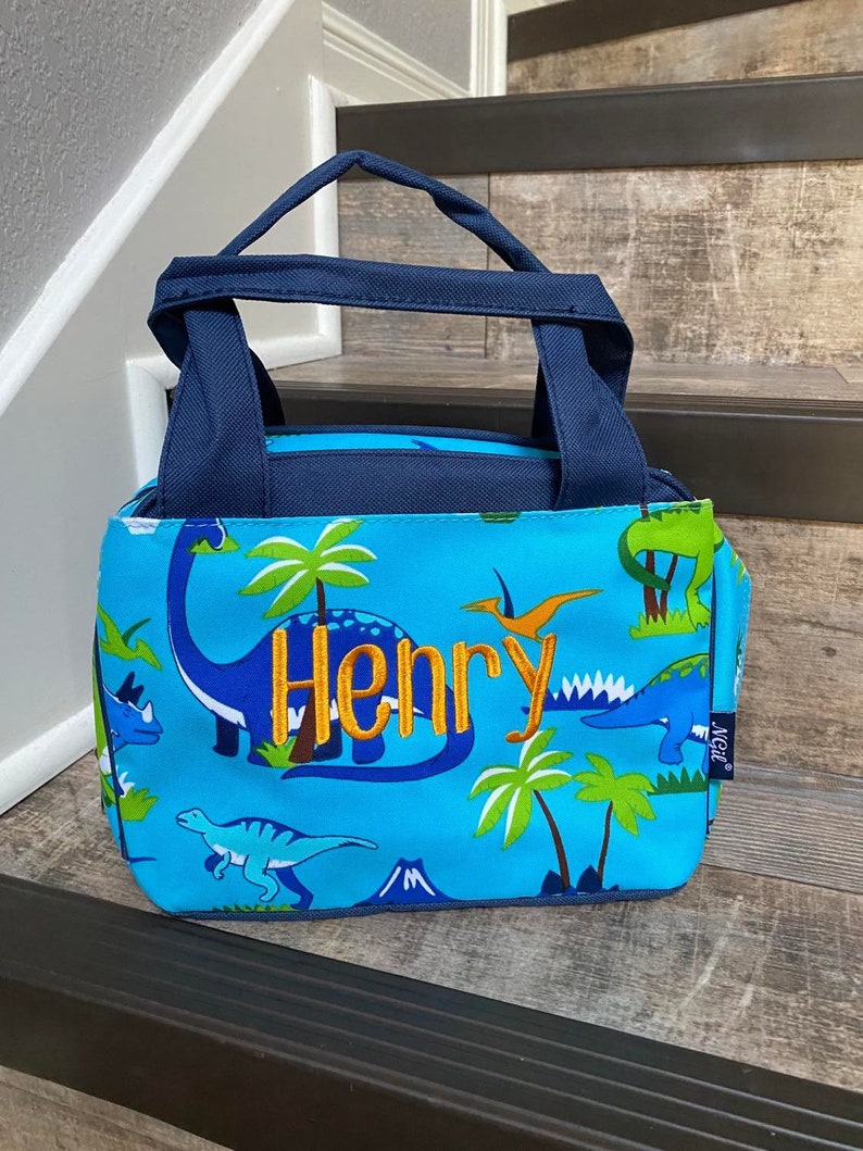 May include: Blue lunch bag with a dinosaur print and the name "Henry" embroidered in orange. The bag has a navy blue handle and a blue and green lining.