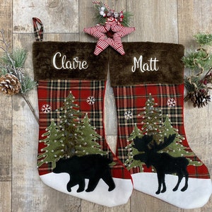 May include: Two personalized Christmas stockings with a plaid design. The stocking on the left has a brown bear and the stocking on the right has a moose. The stockings are decorated with green trees, snowflakes, and white lights. The stocking on the left is personalized with the name "Claire" and the stocking on the right is personalized with the name "Matt".