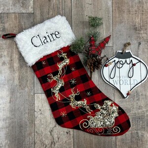 May include: A Christmas stocking with a red and black buffalo plaid pattern, embroidered with a gold Santa and reindeer design. The stocking has a white faux fur cuff with the name "Claire" embroidered in black. A white ornament with the words "Joy to the World" is also present.