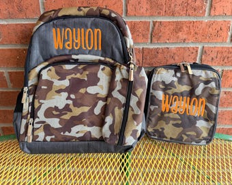 May include: A backpack and a matching lunch bag featuring a camouflage pattern in shades of brown, tan, and green. Both items have the name Waylon embroidered in bright orange thread. The backpack has a dark gray top section and metal zippers.
