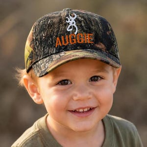 May include: A camouflage baseball cap featuring a white embroidered buck logo and the name AUGGIE stitched in orange thread on the front panel. The cap has a structured crown and a curved brim, displaying a woodland pattern of green and brown.