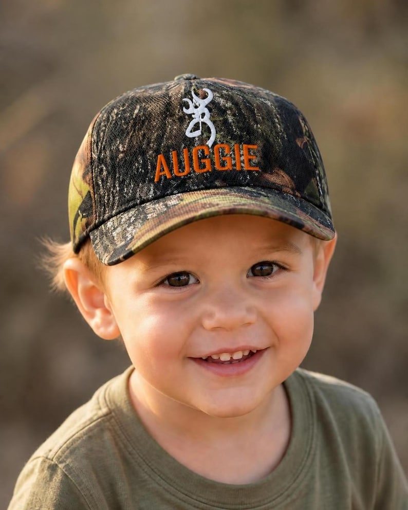 May include: A camouflage baseball cap featuring a white embroidered buck logo and the name AUGGIE stitched in orange thread on the front panel. The cap has a structured crown and a curved brim, displaying a woodland pattern of green and brown.