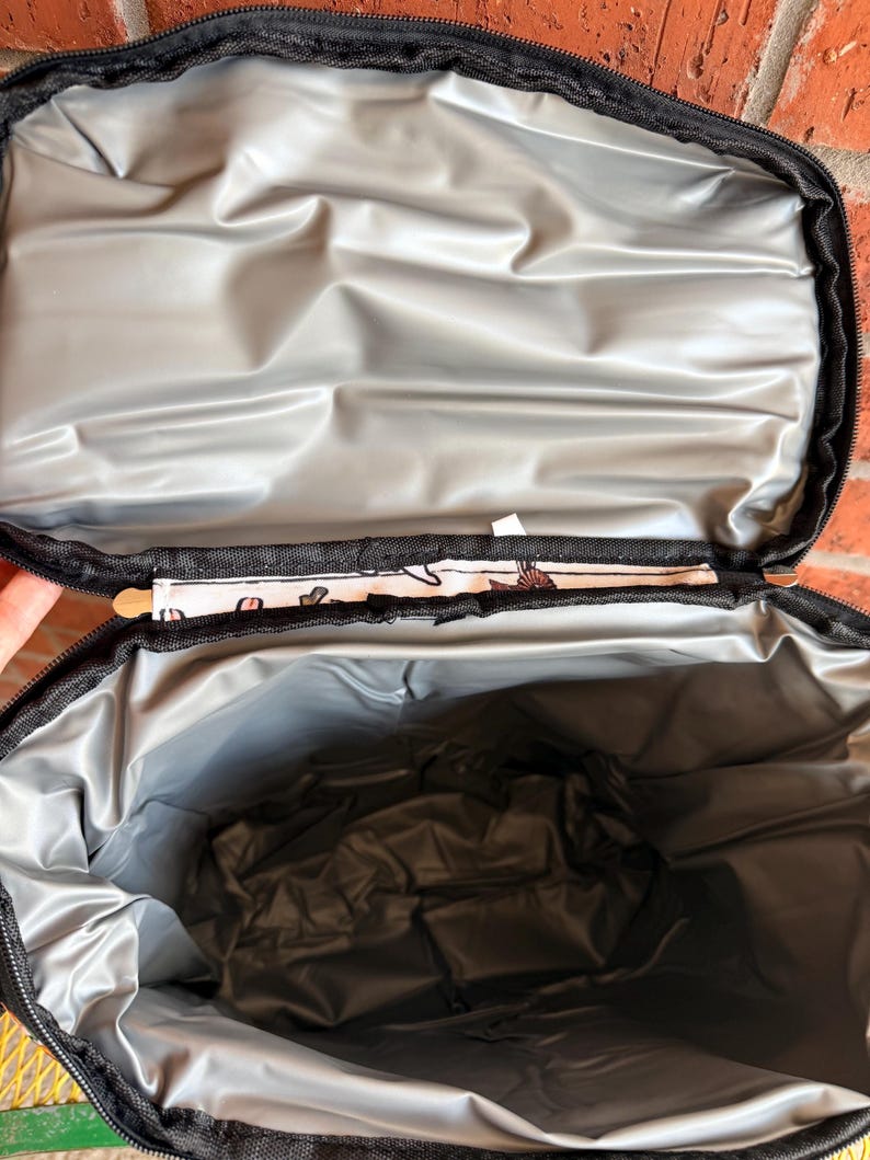 Puede incluir: Bolsa de almuerzo aislante negra abierta con interior plateado. La bolsa tiene una cremallera negra y un ribete estampado. El interior es de un material plateado reflectante, dise&ntilde;ado para mantener el contenido fr&iacute;o o caliente. La bolsa est&aacute; abierta, revelando el espacioso interior.