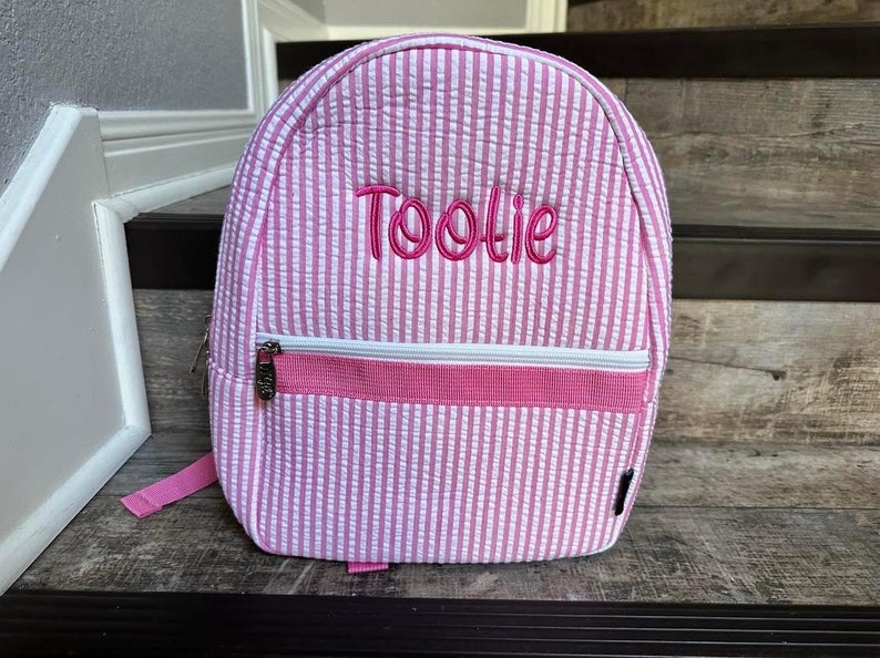 May include: Pink and white striped backpack with the name "Toolie" embroidered in pink. The backpack has a zippered front pocket with a white zipper and a pink strap. The bag is sitting on a wooden surface.