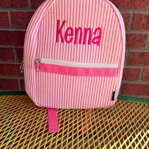 May include: Pink and white striped backpack with the name "Kenna" embroidered in pink. The backpack has a front zipper pocket, a top zipper, and a pink handle. The backpack is sitting on a yellow metal surface.