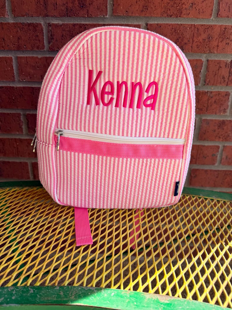 May include: Pink and white striped backpack with the name "Kenna" embroidered in pink. The backpack has a front zipper pocket, a top zipper, and a pink handle. The backpack is sitting on a yellow metal surface.