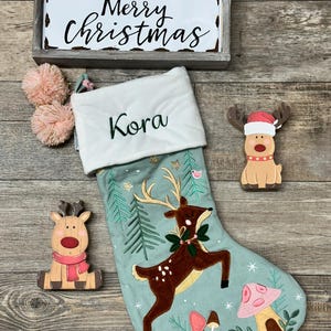 May include: A light green Christmas stocking with a white cuff embroidered with the name Kora, featuring a brown deer, pine trees, and mushrooms. Includes a wooden Merry Christmas sign and two wooden reindeer ornaments with pom-poms.