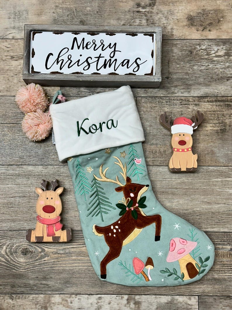May include: A light green Christmas stocking with a white cuff embroidered with the name Kora, featuring a brown deer, pine trees, and mushrooms. Includes a wooden Merry Christmas sign and two wooden reindeer ornaments with pom-poms.