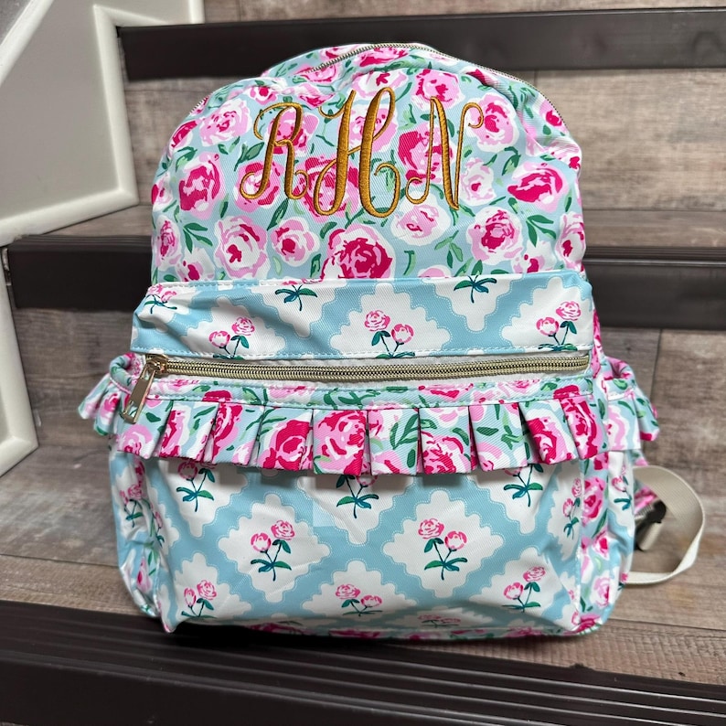 May include: A floral backpack with a light blue background and pink roses. The backpack has a gold zipper and a ruffle detail on the front pocket. Gold embroidered initials are on the top of the backpack.