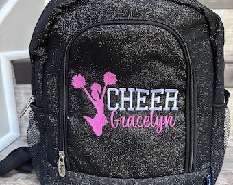 Könnte beinhalten: Schwarzer Glitzer-Rucksack mit einer Vordertasche und seitlichen Netztaschen. Der Rucksack zeigt das Wort "CHEER" in Weiß, eine rosa Cheerleader-Grafik und den Namen "Gracelyn" in rosa Schrift.