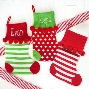 May include: Three personalized Christmas stockings with pom-poms. The stockings are red and green with white stripes and white polka dots. The names "Evan", "Reagan", and "[unclear name]" are embroidered on the stockings.