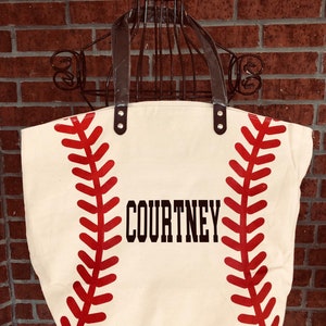 K&ouml;nnte beinhalten: Eine wei&szlig;e Canvas-Tragetasche mit roten Baseballn&auml;hten und dem Namen "COURTNEY" in Schwarz gedruckt.