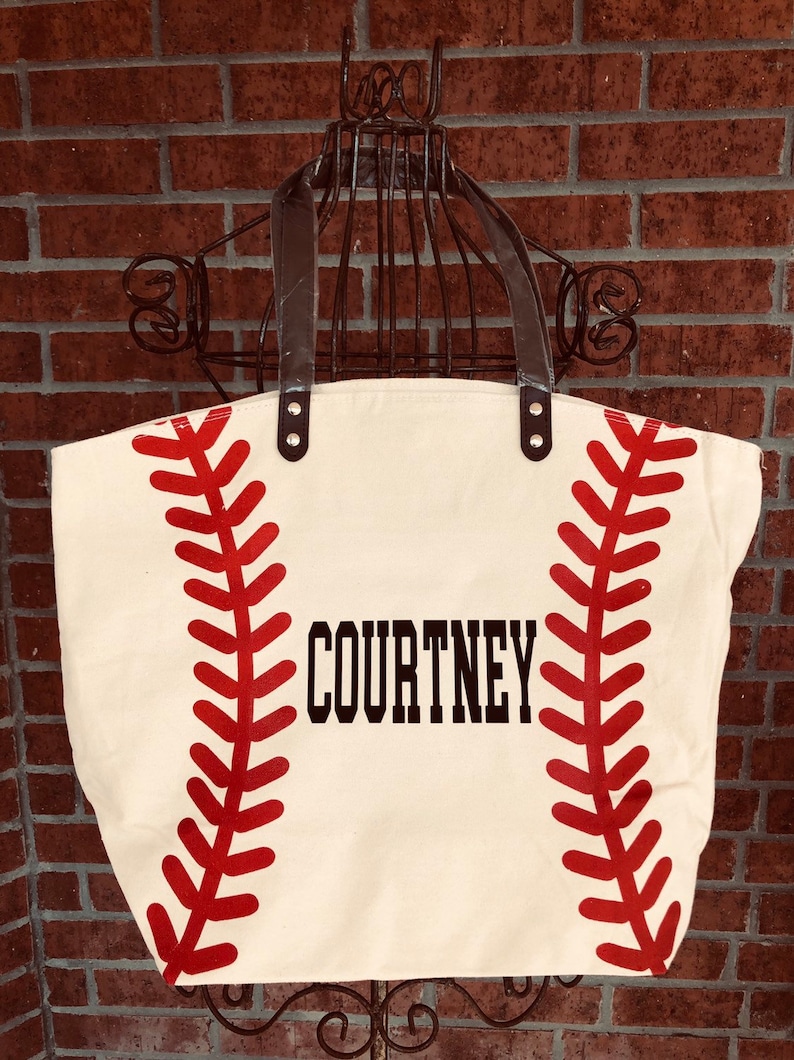 K&ouml;nnte beinhalten: Eine wei&szlig;e Canvas-Tragetasche mit roten Baseballn&auml;hten und dem Namen "COURTNEY" in Schwarz gedruckt.