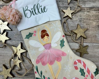 May include: A beige Christmas stocking with a white cuff embroidered with the name Billie in green. The stocking features a fairy in a pink tutu, a candy cane, a peppermint candy, holly leaves, and snowflakes. A pink pom-pom hangs from the cuff.