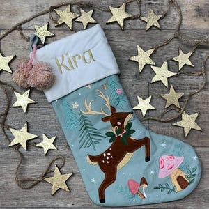 May include: A light blue Christmas stocking featuring an embroidered brown deer with antlers, a green wreath, and pink and brown mushrooms. The white cuff is embroidered with the name Kira in gold thread and includes two pink pom-poms.