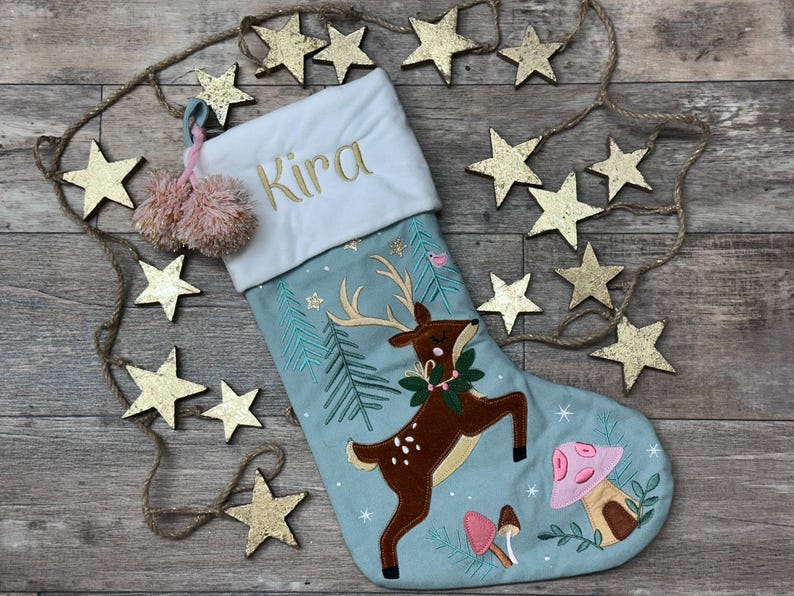 May include: A light blue Christmas stocking featuring an embroidered brown deer with antlers, a green wreath, and pink and brown mushrooms. The white cuff is embroidered with the name Kira in gold thread and includes two pink pom-poms.