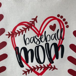 May include: A white canvas tote bag with a red heart design. The heart has a baseball stitch design and the words "baseball mom" in black lettering.