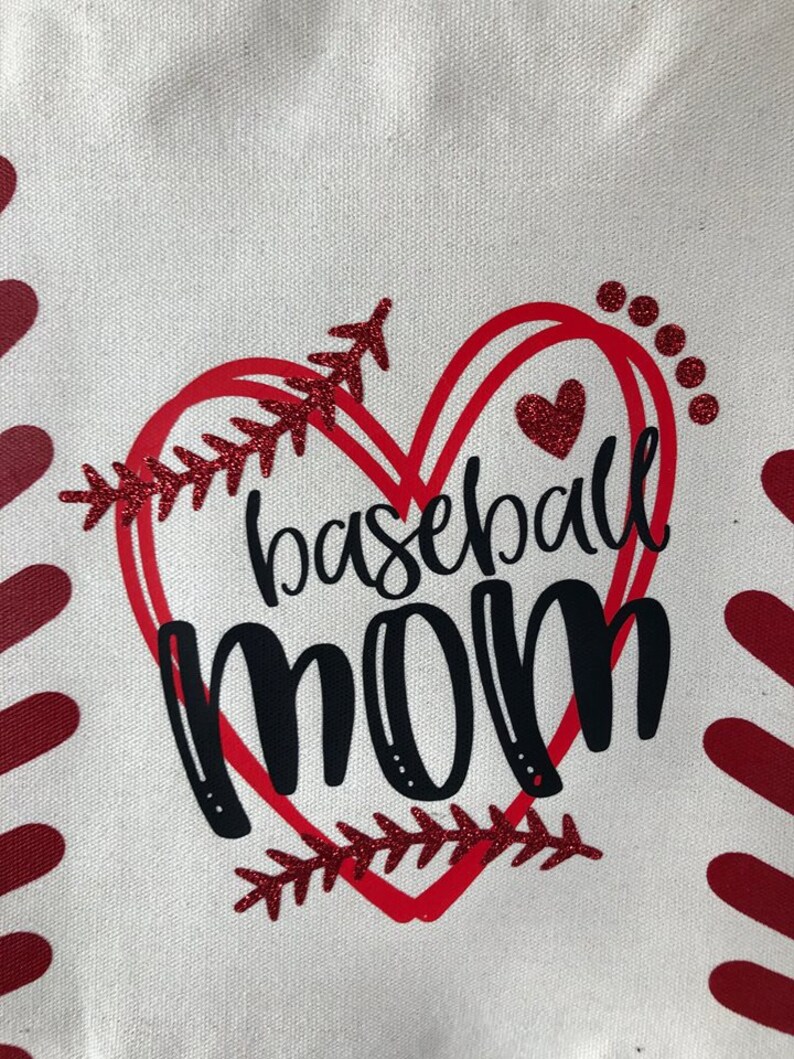 May include: A white canvas tote bag with a red heart design. The heart has a baseball stitch design and the words "baseball mom" in black lettering.