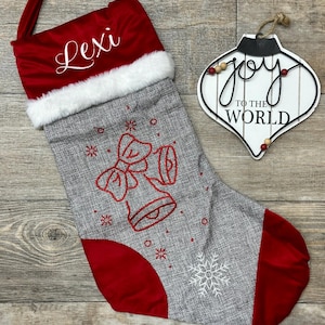 May include: Red and gray Christmas stocking with the name "Lexi" embroidered on the cuff. The stocking features a gray felt body with a red felt cuff and toe. The stocking is decorated with a white snowflake and a red bell with a bow.