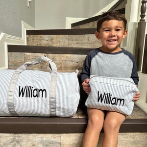 May include: A child sits on stairs holding a small, gray and white striped zippered pouch with the name "William" embroidered on it.  A larger, gray and white striped duffel bag with the name "William" embroidered on it sits on the stairs behind the child.