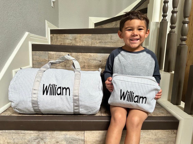 May include: A child sits on stairs holding a small, gray and white striped zippered pouch with the name "William" embroidered on it.  A larger, gray and white striped duffel bag with the name "William" embroidered on it sits on the stairs behind the child.