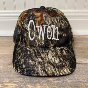 May include: A camouflage baseball cap with the name "Owen" embroidered in white on the front.