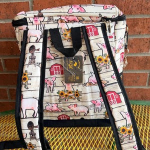 Puede incluir: Mochila con tem&aacute;tica de animales de granja. La mochila presenta un estampado de cerdos rosas, graneros rojos, girasoles y molinos de viento sobre un fondo blanco. Tiene correas negras y una cremallera negra.