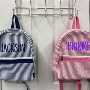 May include: Two small backpacks, one blue and white striped with "JACKSON" embroidered, the other pink and white striped with "BROOKE" embroidered. Both have a zippered front pocket and are hanging on a white door.