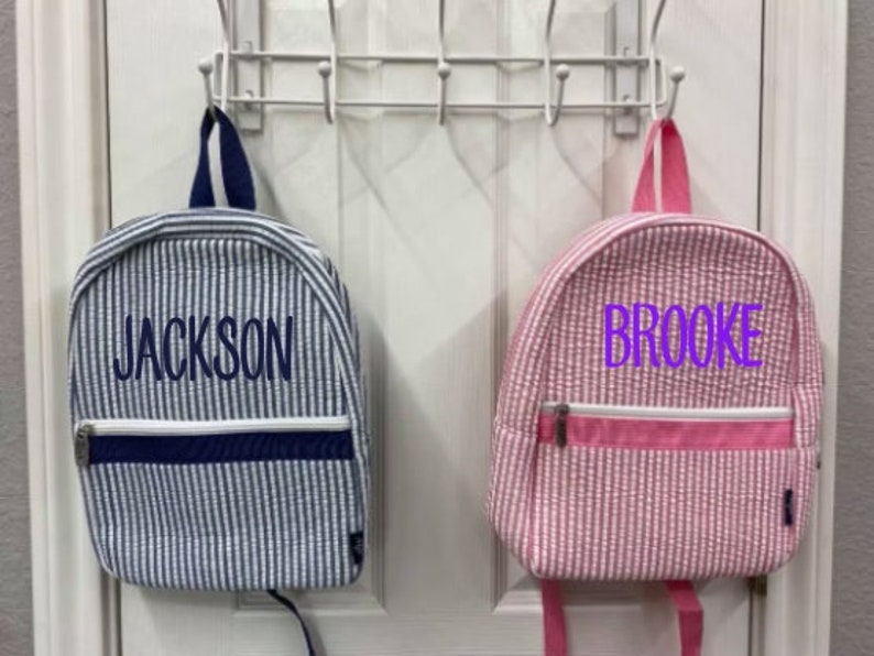 May include: Two small backpacks, one blue and white striped with "JACKSON" embroidered, the other pink and white striped with "BROOKE" embroidered. Both have a zippered front pocket and are hanging on a white door.
