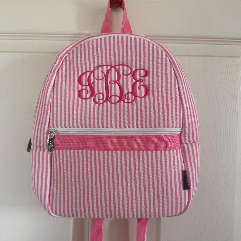 May include: Pink and white striped backpack with a pink embroidered monogram. The backpack has a zippered front pocket, a top handle, and adjustable shoulder straps. The bag is made of a textured fabric.