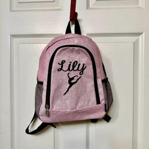 May include: Pink glitter backpack with black trim and a black silhouette of a dancer. The backpack has a zippered pocket and a mesh pocket on the side. The name "Lily" is embroidered on the front of the backpack.