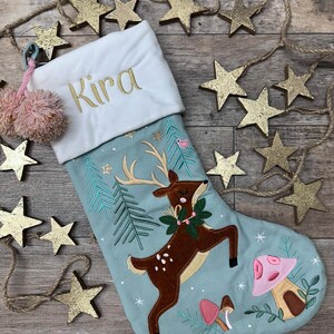May include: A light blue Christmas stocking with a white cuff embroidered with the name Kira in gold thread. The stocking features a brown deer with antlers, green pine trees, and pink mushrooms. A pink pom-pom tassel hangs from the top corner.