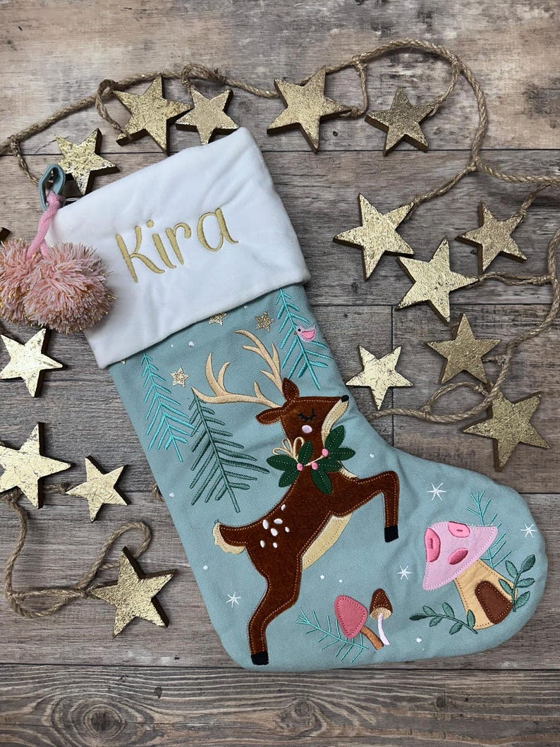 May include: A light blue Christmas stocking with a white cuff embroidered with the name Kira in gold thread. The stocking features a brown deer with antlers, green pine trees, and pink mushrooms. A pink pom-pom tassel hangs from the top corner.