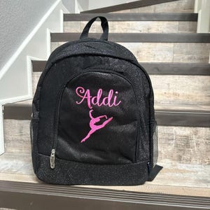May include: Black glitter backpack with a pink embroidered dancer and the name "Addi" on the front.
