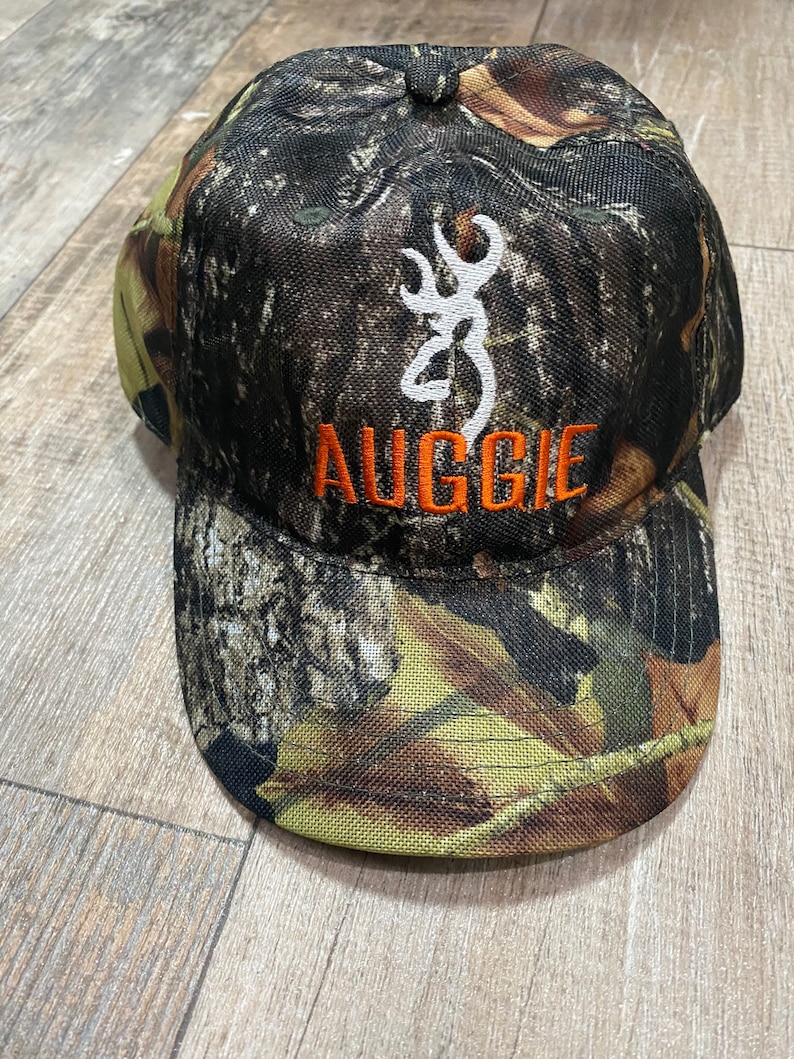 May include: Camouflage baseball cap with the word "AUGGIE" in orange lettering. The cap features a white emblem of a deer. The hat is made of a durable fabric and is designed for outdoor activities.