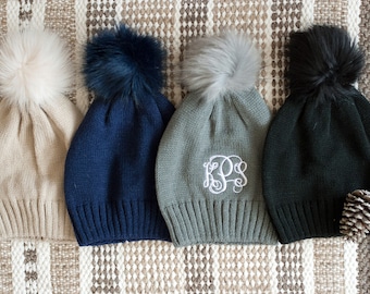 May include: Four knit winter hats with faux fur pom poms. The hats are in beige, navy blue, gray, and black. The gray hat has a white monogram "KPS" embroidered on the front.