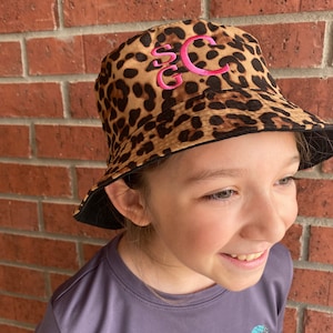 May include: A brown and black leopard print bucket hat with a pink embroidered monogram "SC".
